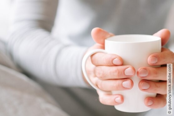 Frau hält weißen Becher in der Hand und wärmt ihre Hände daran.